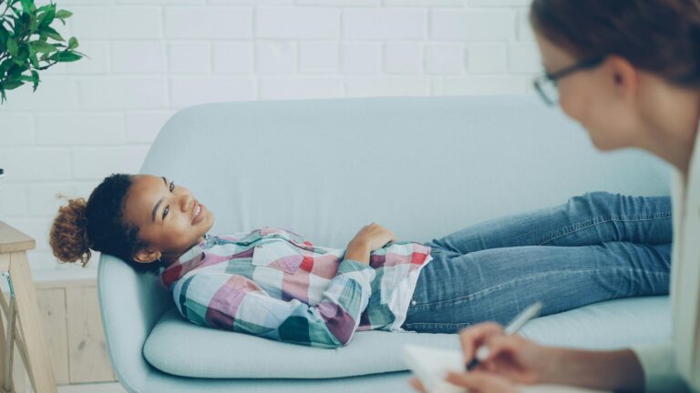 Resources A woman in therapy, smiling while lying on a couch, with a counselor taking notes.